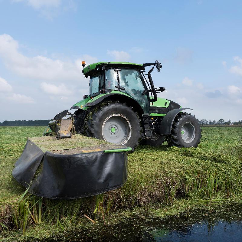 Een zware groene trekker met een maaimachine maait het gras aan een slootkant.